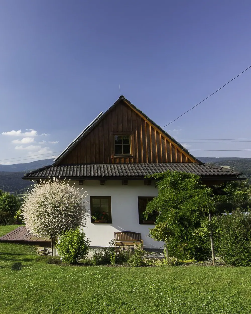 Dům s dřevěným štítem na kraji louky, vlevo dřevěná houpačka se skluzavkou, za domem pásmo kopců.