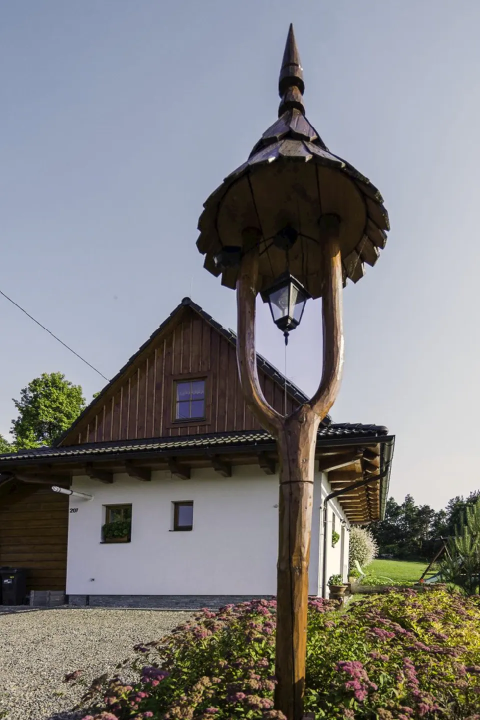 Vyřezávaná dřevěná lucerna se stříškou na sloupu před domem, za ní bílý dům s dřevěným štítem a v popředí kvetoucí keře.