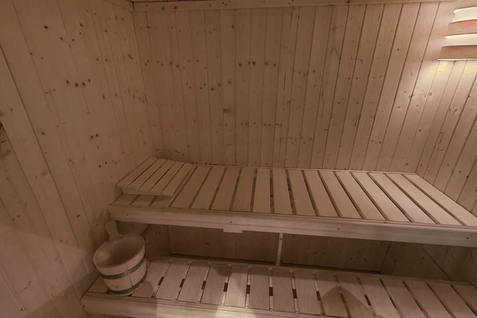Finská sauna v koupelně zevnitř: lavice ve dvou výškách z dřevěných latí, dřevěné vědro a lampa na stěně z palubek.