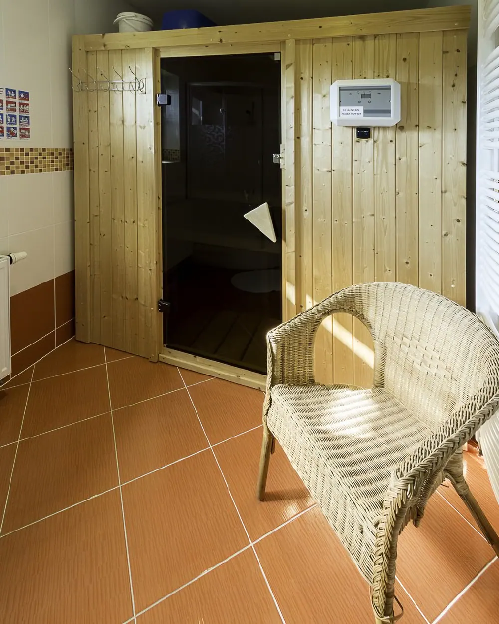 Dřevěná kabina finské sauny se skleněnými dveřmi a ovládacím panelem, před ní proutěné křeslo na dlažbě koupelny.