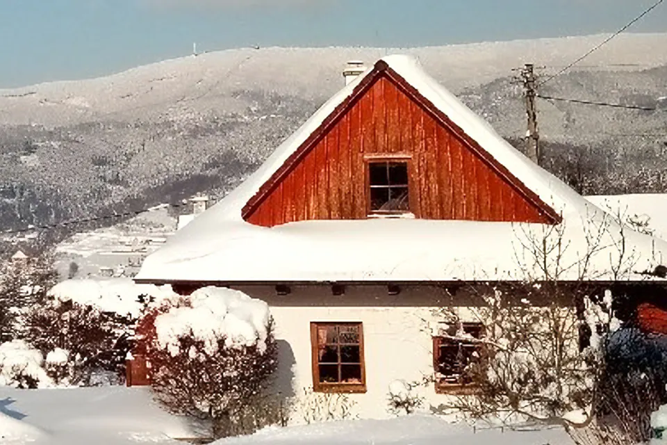 Dům s červeným dřevěným štítem pod sněhem, za ním přes údolí zasněžený hřeben pod modrým nebem.