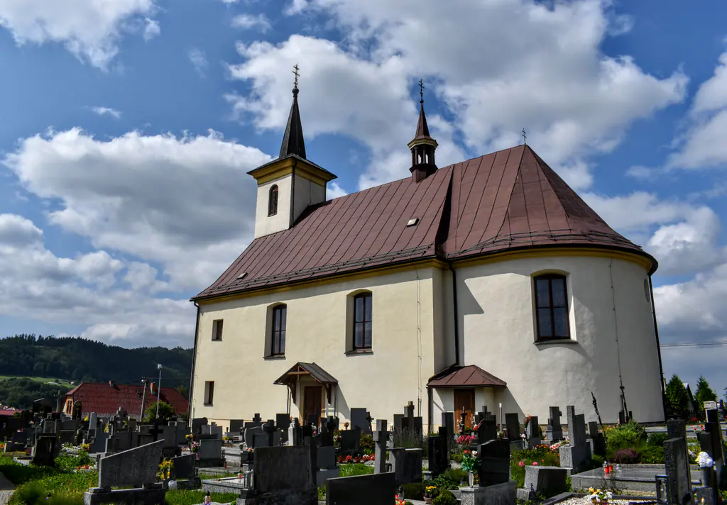 Bílý kostel sv. Josefa v Hutisku s věží a hnědou střechou, před ním hřbitov, nad ním letní mraky