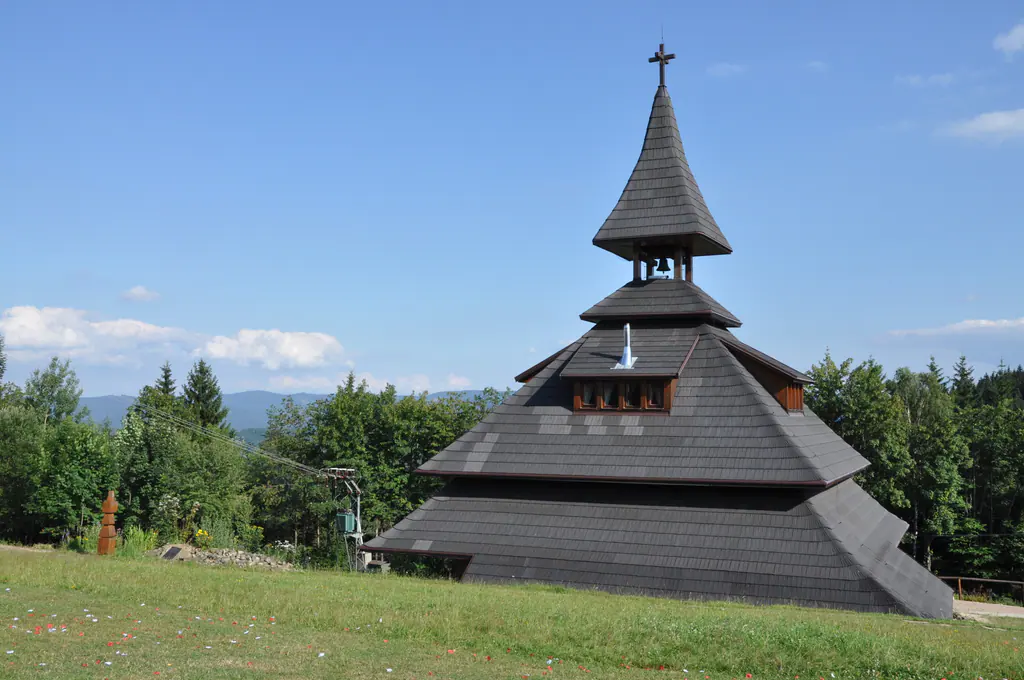 Dřevěná zvonice Soláň se stupňovitou šindelovou střechou a křížem na louce, za ní smrky a hřebeny.