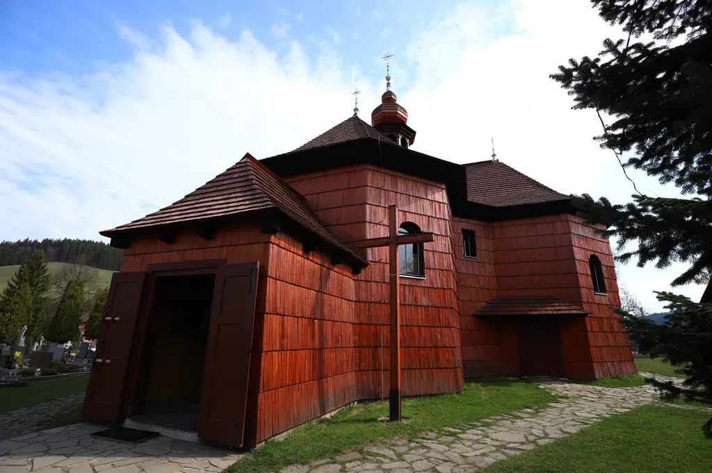 Dřevěný kostel Panny Marie Sněžné obložený červenohnědými šindeli, před ním dřevěný kříž a kamenná cesta