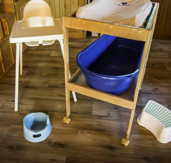 Bílá krmicí židlička vedle dřevěného přebalovacího pultu na kolečkách, pod jeho deskou modrá dětská vanička, na podlaze nočník a stupínek