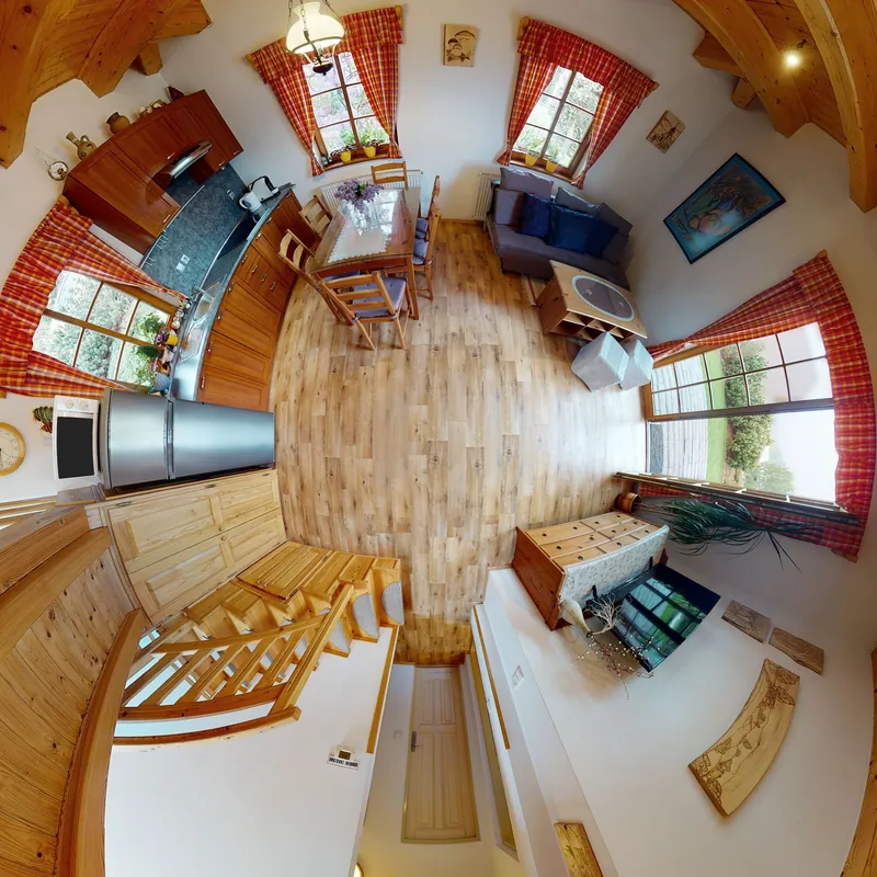 Obývák s kuchyní jako jeden kulatý obraz shora, fotka 360°: borový trámový strop kolem dokola, kuchyňská linka, jídelní stůl, pohovka s modrými polštáři, schodiště do podkroví a okna s károvanými záclonami.