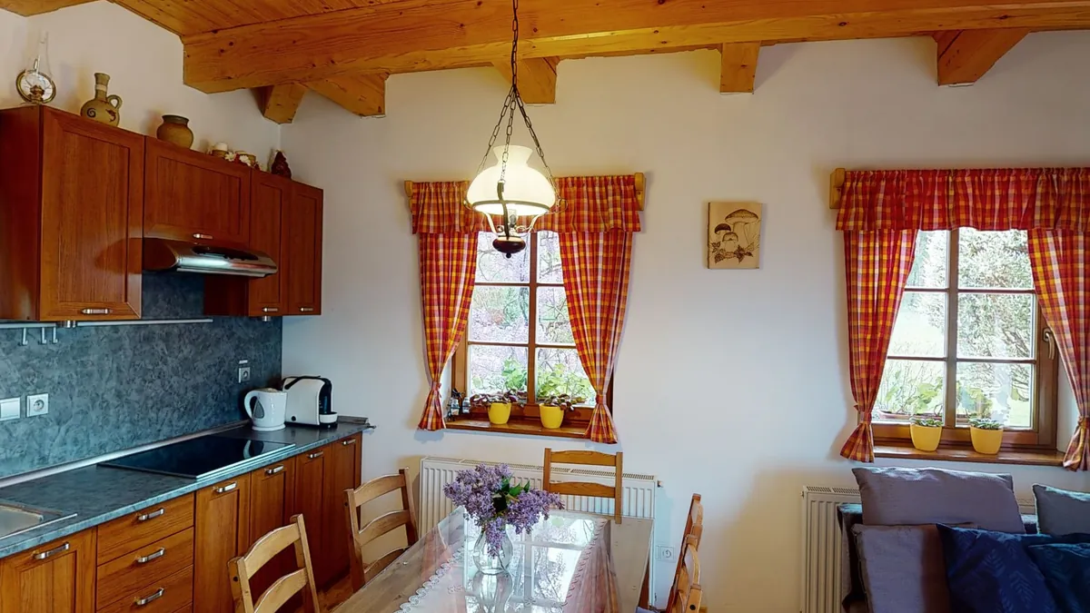 Obývák s kuchyní z výšky očí: borový trámový strop, dřevěná kuchyňská linka s modrošedým obkladem, dvě okna s károvanými záclonami, lampa nad jídelním stolem s kyticí šeříku a pohovka s modrými polštáři.